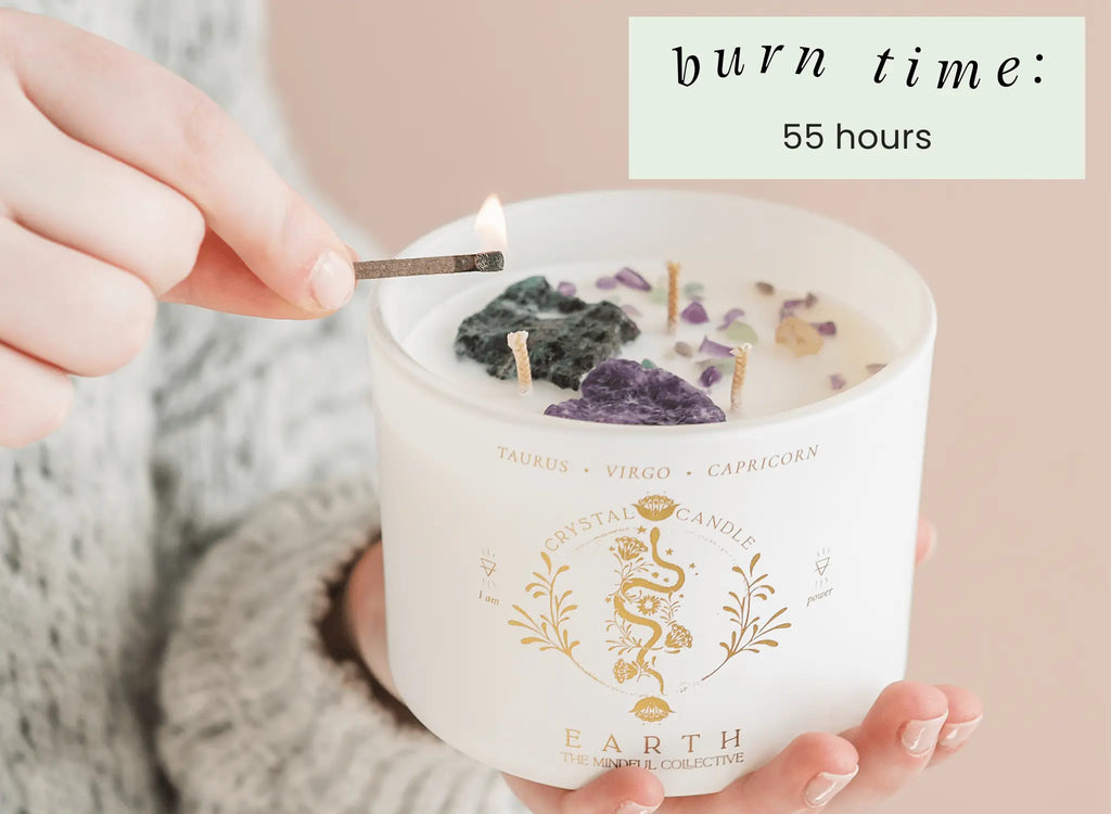 A person lights the Earth Elements Candle 12oz by THE MINDFUL COLLECTIVE, featuring stones inside. The label lists Taurus, Virgo, Capricorn. Burn time: 55 hours—a grounding candle perfect for Earth Signs.