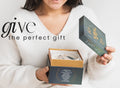 A woman in a white sweater opens a dark blue gift box containing THE MINDFUL COLLECTIVE Earth Elements Candle 12oz. Text on the image reads, give the perfect gift.