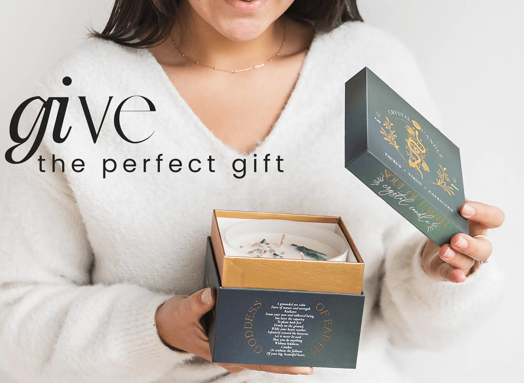 A woman in a white sweater opens a dark blue gift box containing THE MINDFUL COLLECTIVE Earth Elements Candle 12oz. Text on the image reads, give the perfect gift.