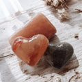 Two polished heart stones—orange and dark gray—rest beside Modern Skyn Alchemy LLC.’s Exfoliating Salt Stones: Pink Bar on a light wood surface with dried white flowers, creating a serene setting perfect for your wellness rituals.