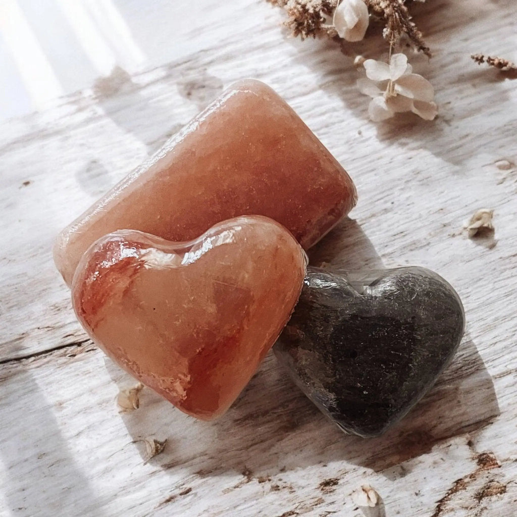 Two polished heart stones—orange and dark gray—rest beside Modern Skyn Alchemy LLC.’s Exfoliating Salt Stones: Pink Bar on a light wood surface with dried white flowers, creating a serene setting perfect for your wellness rituals.