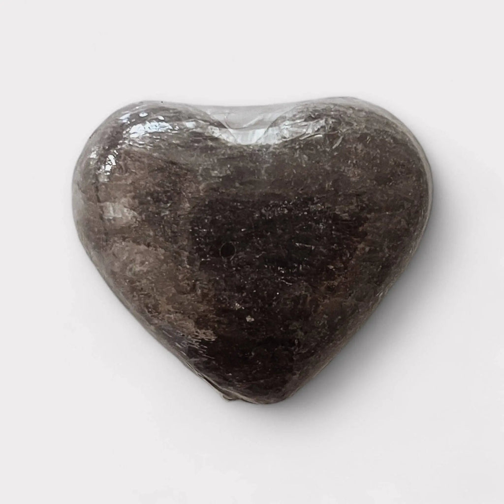 The Exfoliating Salt Stones: Pink Bar by Modern Skyn Alchemy LLC. is a heart-shaped, dark stone wrapped in clear plastic—perfect for upgrading your wellness routine—shown on a plain white background.