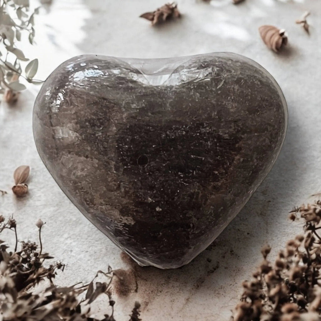 A heart-shaped, pink Exfoliating Salt Stone by Modern Skyn Alchemy LLC. rests on a light surface amid dried flowers and foliage, creating a rustic vibe—perfect inspiration for your wellness routine.