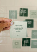 A hand with light purple nails holds a JaxKelly Shower Affirmation™ Cards - Abundance in the shower, reading: I am okay with saying no to what does not serve me. More waterproof cards are blurred in the tiled background.