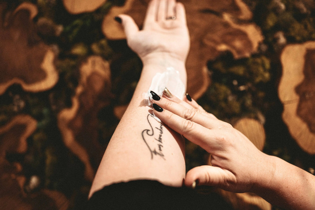 A person applies Salt & Sage Apothecary’s I AM Abundant Whipped Organic Body Butter to their forearm, displaying a cursive tattoo. Their nails are painted black and the background features patterned wooden surfaces.