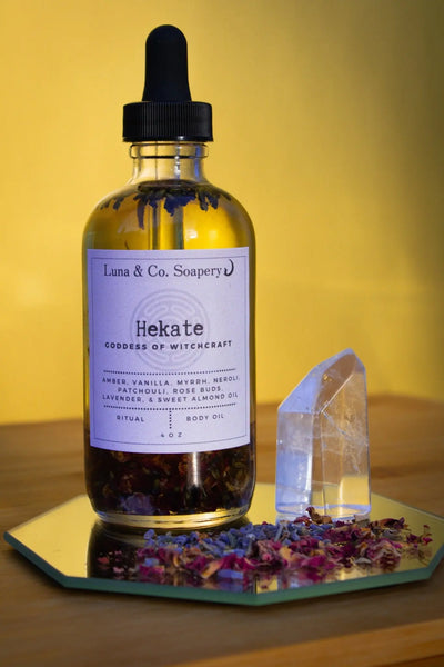 A bottle of Hekate Body Oil by Luna & Co. Soapery rests on a mirror with two clear crystals and dried flower petals, glowing against a warm yellow background—a nod to goddess body oil rituals.