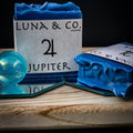 A blue Jupiter Soap by Luna & Co. Soapery rests on a wooden tray with a mirror and blue marble, evoking a ritual bath scene against a dark background. Another soap bar lies flat, partially seen.