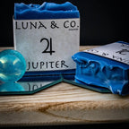A blue Jupiter Soap by Luna & Co. Soapery rests on a wooden tray with a mirror and blue marble, evoking a ritual bath scene against a dark background. Another soap bar lies flat, partially seen.