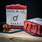 Two reddish-pink Mars Soap bars from Luna & Co. Soapery, each featuring the Mars symbol, sit atop mirrors on wood. A brown crystal cluster nearby enhances ritual bath vibes and planetary energy against a black background.
