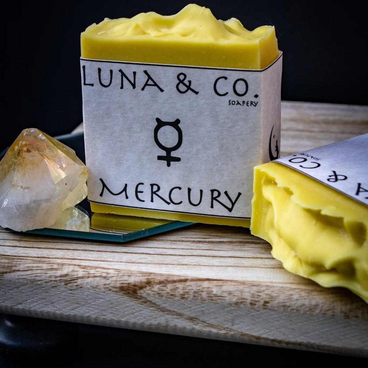The yellow Mercury Soap by Luna & Co. Soapery, perfect for ritual cleansing and planetary magick, rests on a wooden board next to a crystal and another soap, all set against a dark background.
