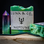 A green Neptune Soap bar from Luna & Co. Soapery featuring a trident symbol sits next to a vibrant crystal on wood, ideal for ritual baths and spiritual healing, set against a dark background.
