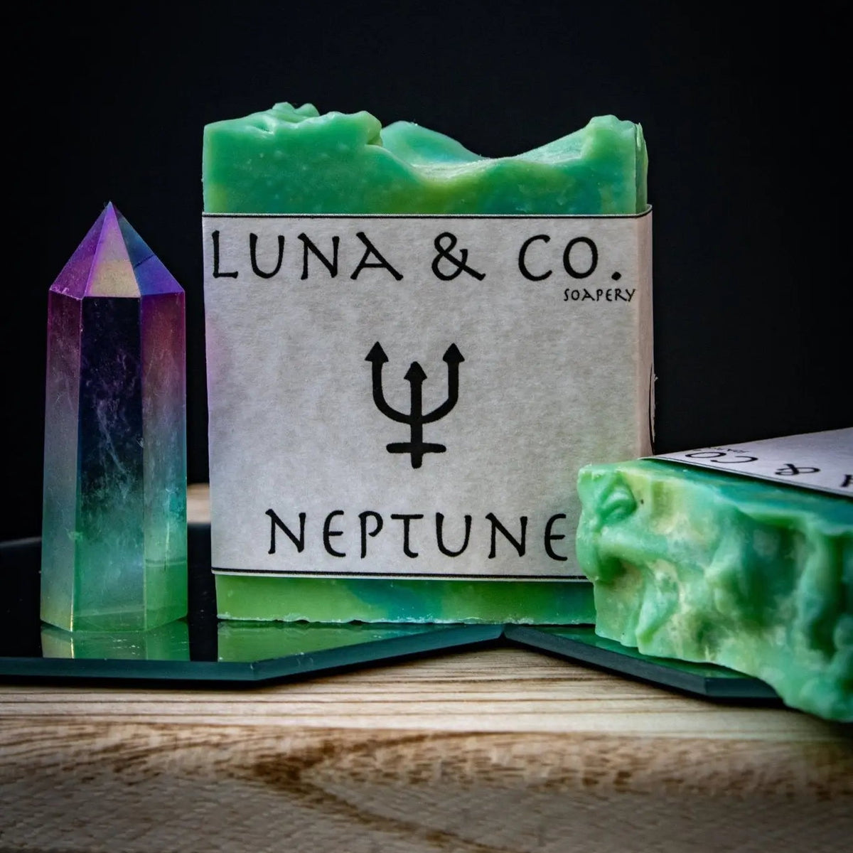 A green Neptune Soap bar from Luna & Co. Soapery featuring a trident symbol sits next to a vibrant crystal on wood, ideal for ritual baths and spiritual healing, set against a dark background.