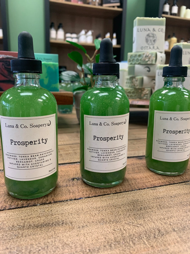 Three glass dropper bottles of Luna & Co. Soaperys Prosperity Ritual Manifestation Oil * New Formula, filled with green liquid and part of the Manifestation Collection, sit on a wooden surface with blurred soaps in the background.