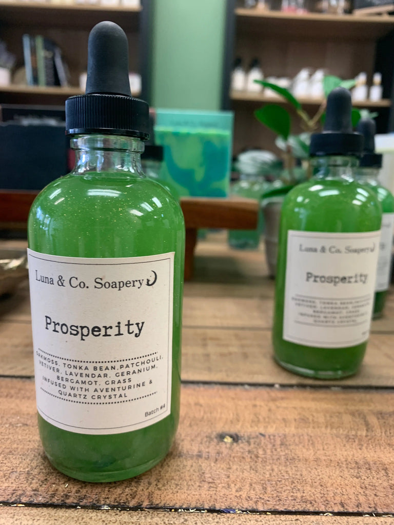 A glass bottle of Luna & Co. Soapery’s Prosperity Ritual Manifestation Oil * New Formula, filled with green liquid and topped with a dropper lid, sits on a wooden table. Part of the Manifestation Collection, another bottle is blurred in the background.