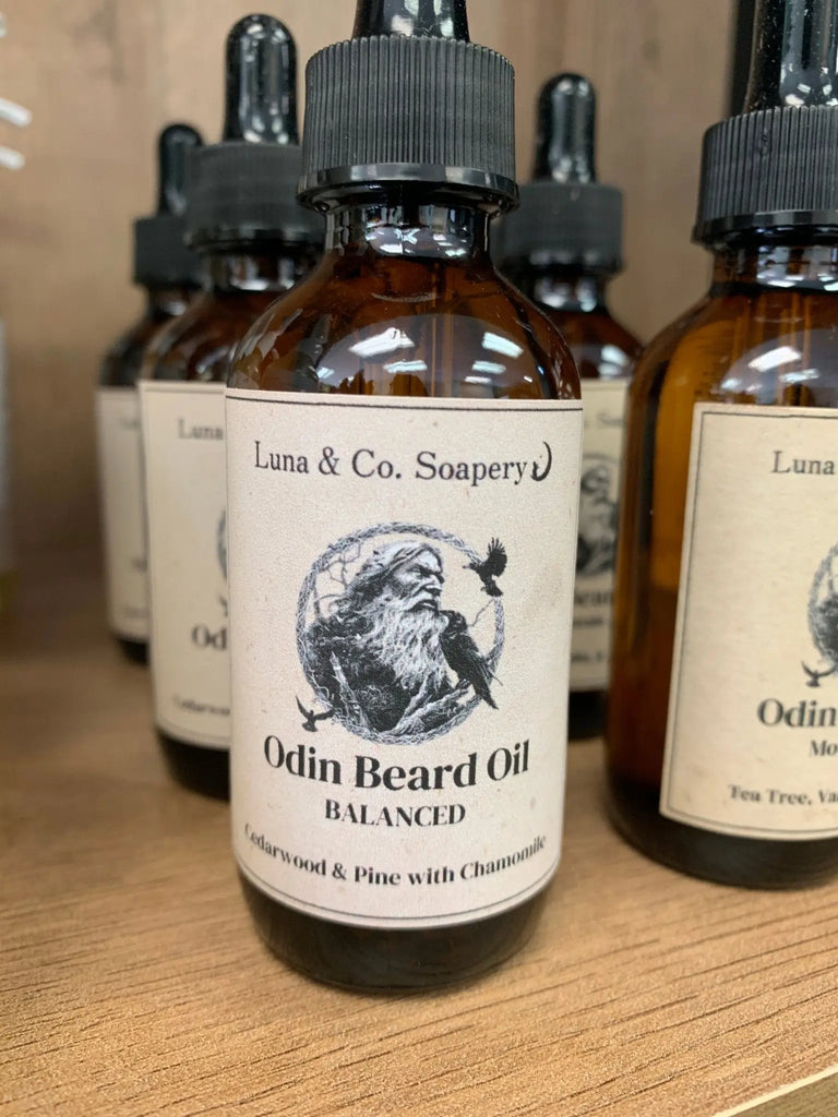 A brown glass dropper bottle labeled Odins Beard Oil from Bath Alliance sits on a wooden shelf. The label features an illustration of Odin and highlights cedarwood, pine, chamomile, and nourishing jojoba oil as ingredients.