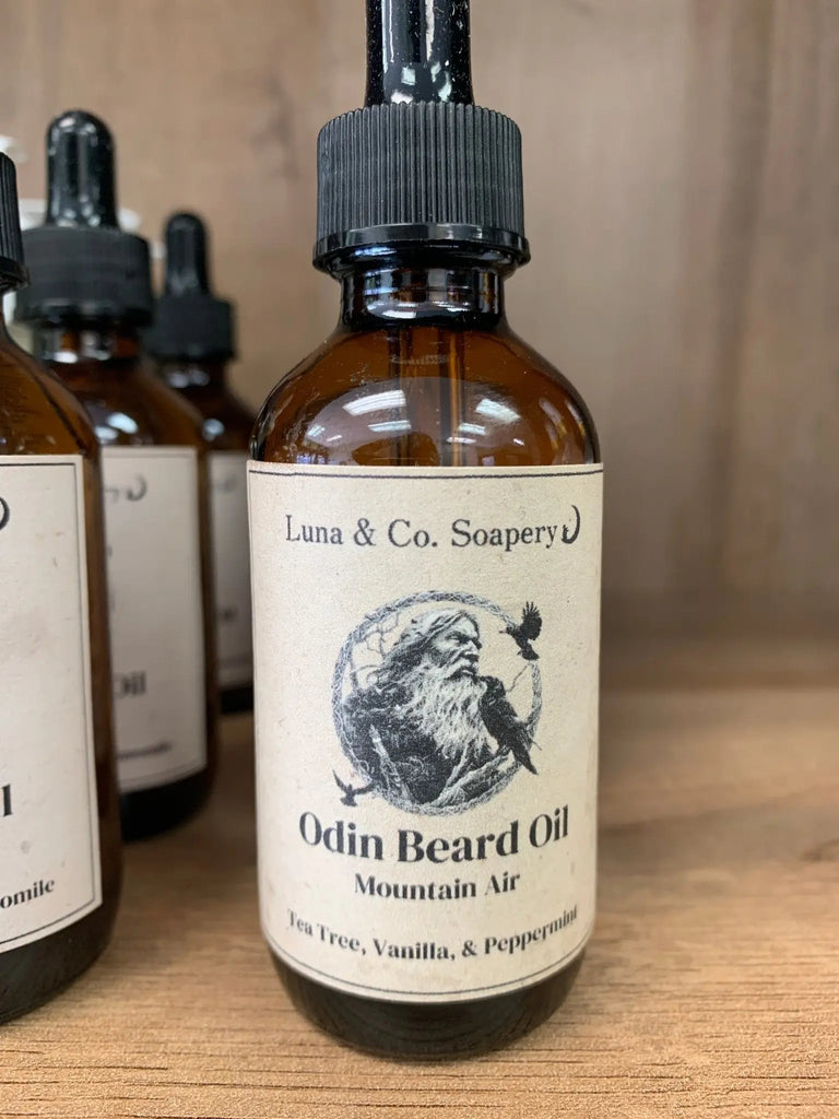 A brown glass dropper bottle labeled Odins Beard Oil by Bath Alliance, featuring an Odin illustration and listing essential oils such as tea tree, vanilla, peppermint, and nourishing jojoba oil.
