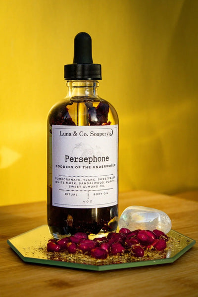 A glass dropper bottle of Persephone Body Oil by Luna & Co. Soapery rests on a mirrored surface with dried rosebuds and a crystal, evoking feminine energy against a bold yellow background.