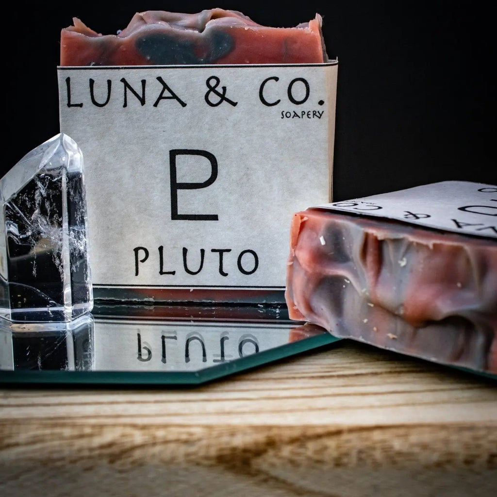 A handcrafted bar of Luna & Co. Soapery Pluto Soap stands upright next to a broken glass shard on a mirror, with another bar lying flat—all set against a dark backdrop, suggesting the mystery of ritual bath and spiritual transformation.