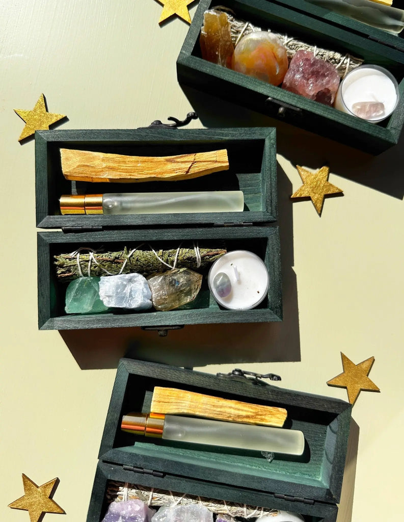 Three PROTECTING MY ENERGY KIT boxes by The 12th Moon, each filled with crystals, herbs, sage, Palo Santo sticks, and glass perfume rollers for “Dream + Manifest” intentions, sit on a yellow surface with gold star cutouts basking in sunlight.