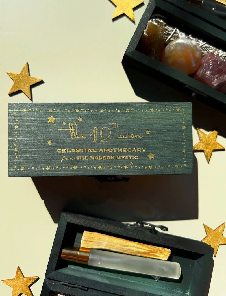 A dark green PROTECTING MY ENERGY KIT by The 12th Moon, labeled “Crystal Kit - Smudge - Sage - Palo: INTENTIONS KIT to Dream + Manifest,” is surrounded by gold paper stars, with perfume vials and stress relief crystals partially visible nearby.