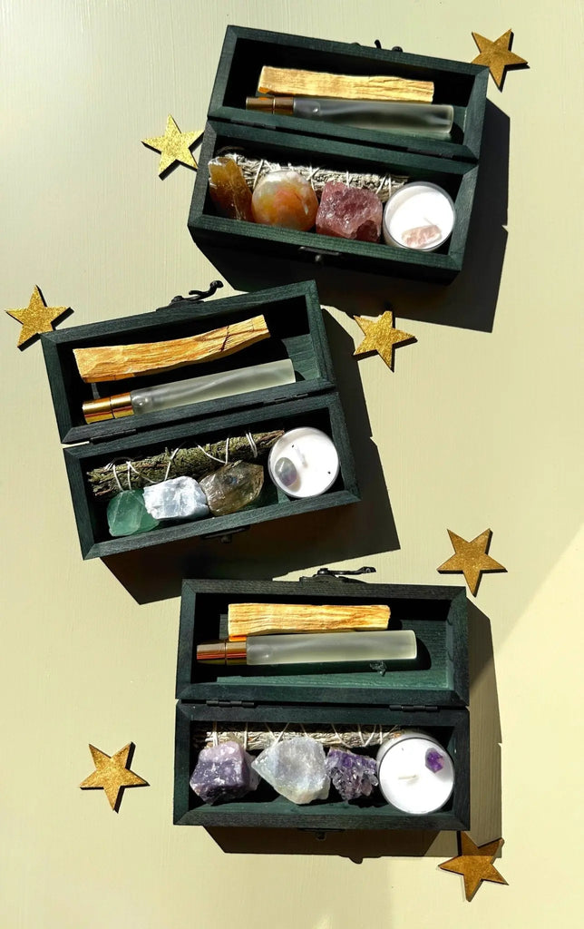 Three PROTECTING MY ENERGY KITS by The 12th Moon—each a crystal kit with stress relief crystals, palo santo, sage, and a white tealight—are arranged diagonally on a light background with gold stars.