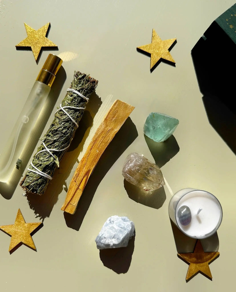 A flat lay displays The 12th Moon PROTECTING MY ENERGY KIT—featuring a perfume roller, sage, palo santo, stress-relief crystals, tealight candle, and gold stars—the perfect VIBING KIT to relieve stress and remove negativity.