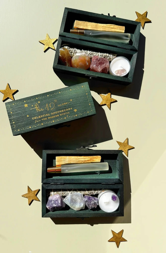 Two green wooden boxes labeled The 12th Moon contain the PROTECTING MY ENERGY KIT—crystals, a small spray bottle, sage, Palo Santo, and a tealight—VIBING KIT to relieve stress and remove negativity, all accented with gold star decorations.