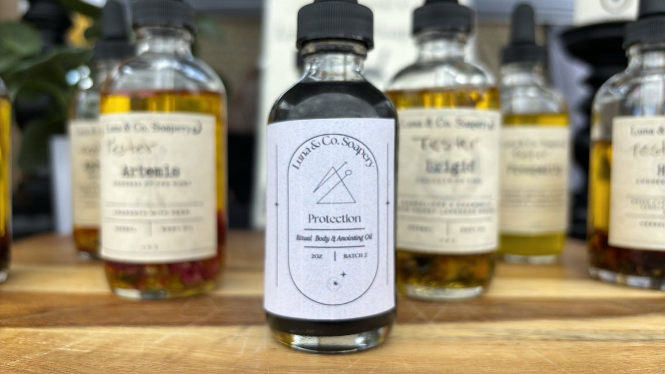 A close-up of a dark glass bottle labeled Saturn Ritual Oil by Luna & Co. Soapery sits on a wooden surface, evoking grounding and protection, with several other labeled bottles softly blurred in the background.