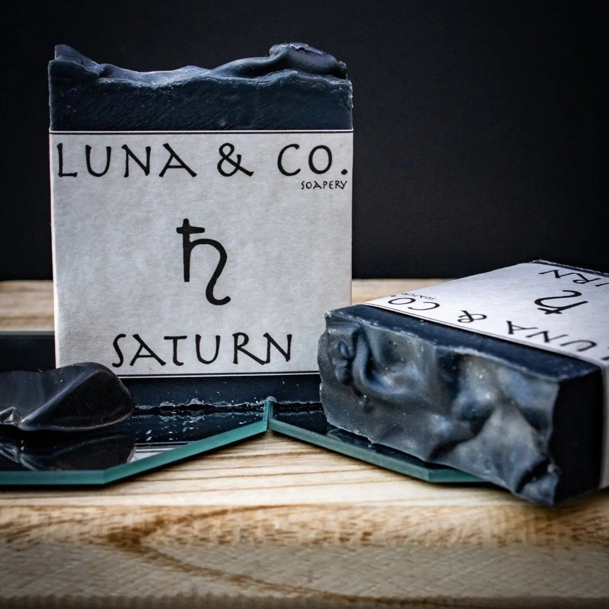 Two black-and-white Saturn Soap bars by Luna & Co. Soapery, crafted with activated charcoal, are arranged on a wooden tray—one upright, one flat—against a dark minimalist backdrop. Elevate your bath ritual with this artisanal touch.