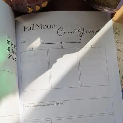 The Soul Care Moon Journal by Bella deLuna is open to a “Full Moon Card Spread” page with three numbered boxes and a question at the bottom. Sunlight and plant shadows cross the page while handwritten notes appear on both sides.
