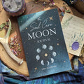 The Soul Care Moon Journal by Bella deLuna rests on a wooden surface with dried flowers, an antler, a wax-sealed letter, a crystal, and colorful cloth—ideal for moon rituals or as your personal manifestation journal.