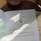 The Soul Care Venus Journal by Bella deLuna features an open Your Venus page with the Venus symbol, fill-in-the-blank prompts, sunlight and shadows on the paper, plus handwritten notes and a candle visible nearby.