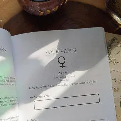 The Soul Care Venus Journal by Bella deLuna features an open Your Venus page with the Venus symbol, fill-in-the-blank prompts, sunlight and shadows on the paper, plus handwritten notes and a candle visible nearby.