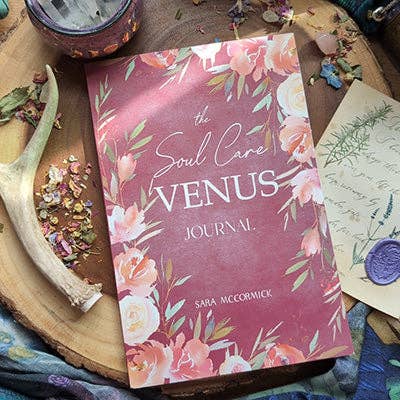The Soul Care Venus Journal by Bella deLuna, featuring a floral cover, sits on a wooden surface amid flower petals, an antler, a candle, and a handwritten letter sealed with purple wax.