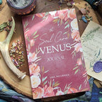 The Soul Care Venus Journal by Bella deLuna, featuring a floral cover, sits on a wooden surface amid flower petals, an antler, a candle, and a handwritten letter sealed with purple wax.
