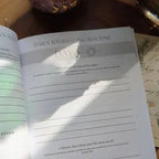 The Bella deLuna Soul Care Venus Journal is open to a “Day 5 Daily Journaling Routine” page with prompts and space to write. Sunlight and shadows play across the journal, with handwritten notes partly shown beside it.