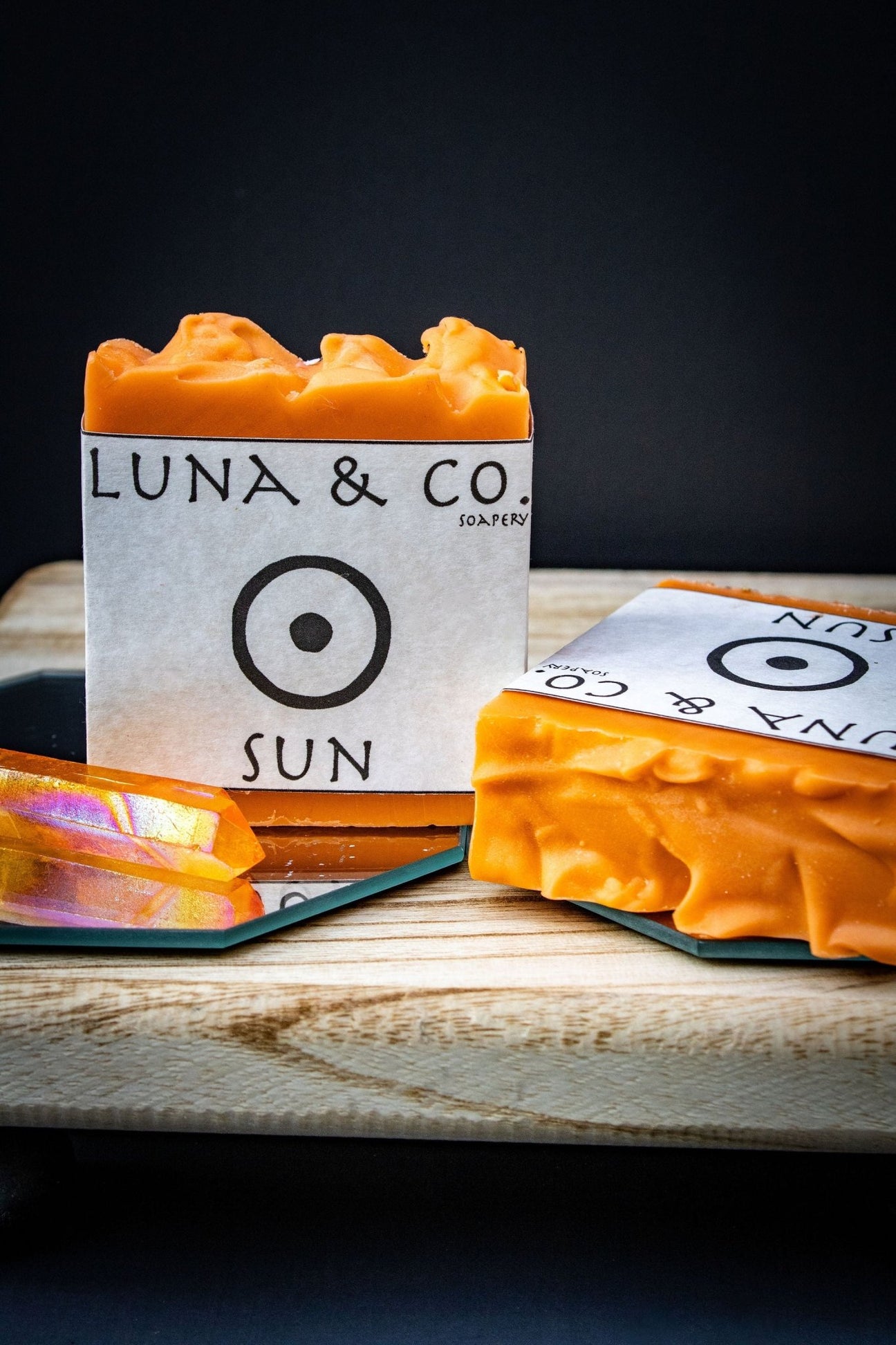 A vibrant orange Sun Soap from Luna & Co. Soapery rests on wood, with a mirror reflecting another bar and an iridescent dish nearby—an ideal addition to your ritual bath against a dark backdrop.