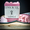 The Venus Soap by Luna & Co. Soapery, a pink bar with a Venus symbol, rests on a wooden board beside rose quartz and another sliced soap—ideal for your ritual bath or manifestation, set against a dark backdrop.